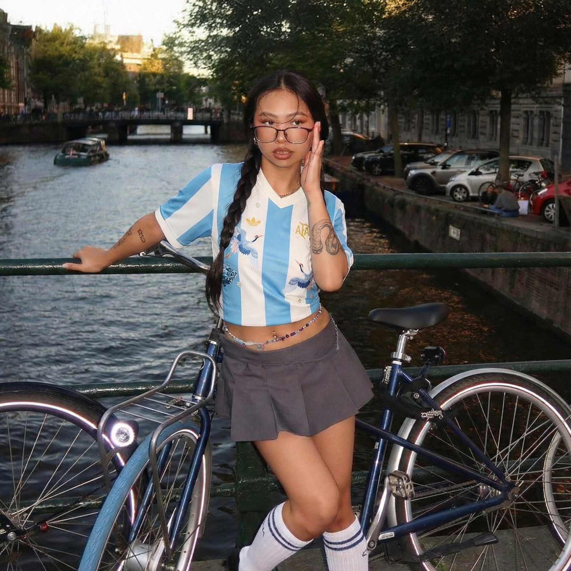 Woman wearing an Argentina home football jersey with sky blue and white stripes, styled as a crop top on a bridge with a bicycle and canal in the background