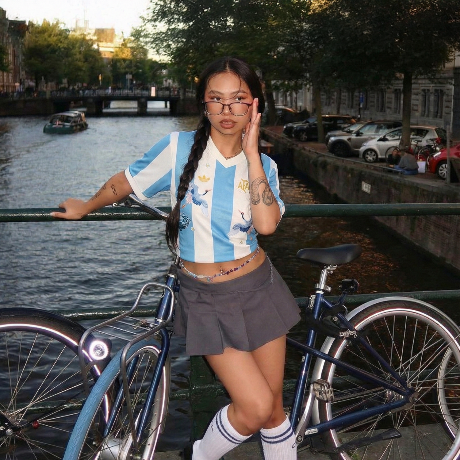 Woman wearing an Argentina home football jersey with sky blue and white stripes, styled as a crop top on a bridge with a bicycle and canal in the background