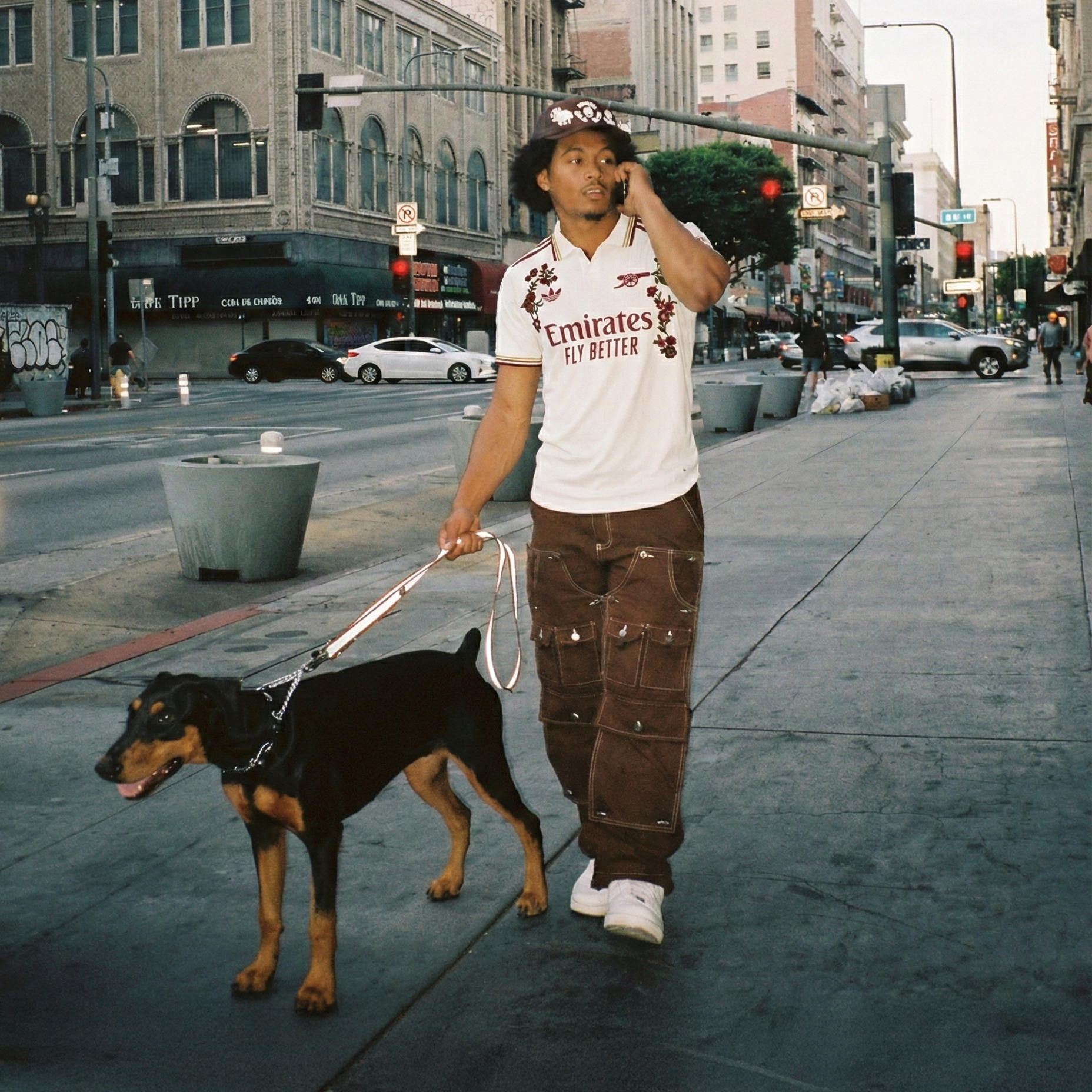 Man wearing Arsenal Embroidered football jersey with red floral embroidery, walking a dog in an urban street setting