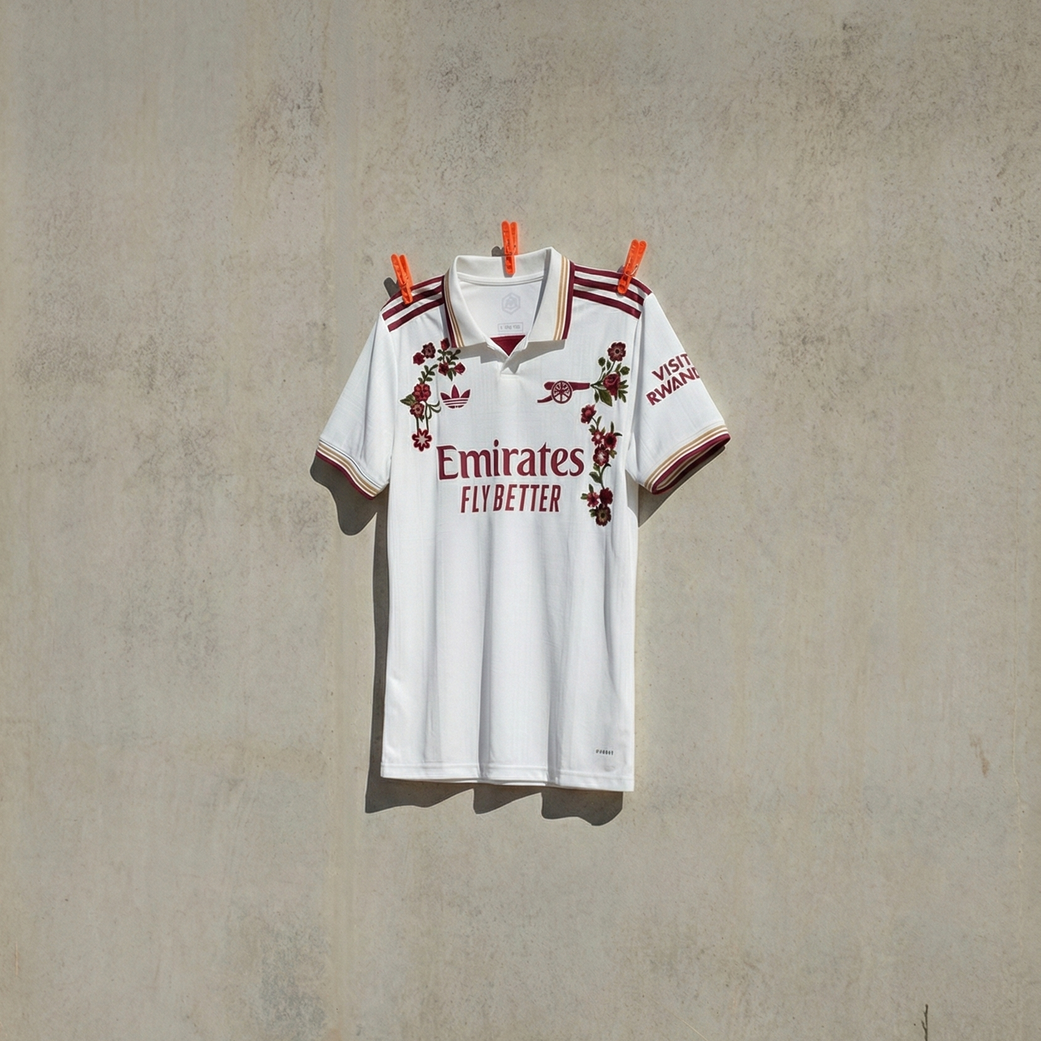Arsenal Embroidered football jersey in white with red floral embroidery, hanging against a concrete wall in natural light