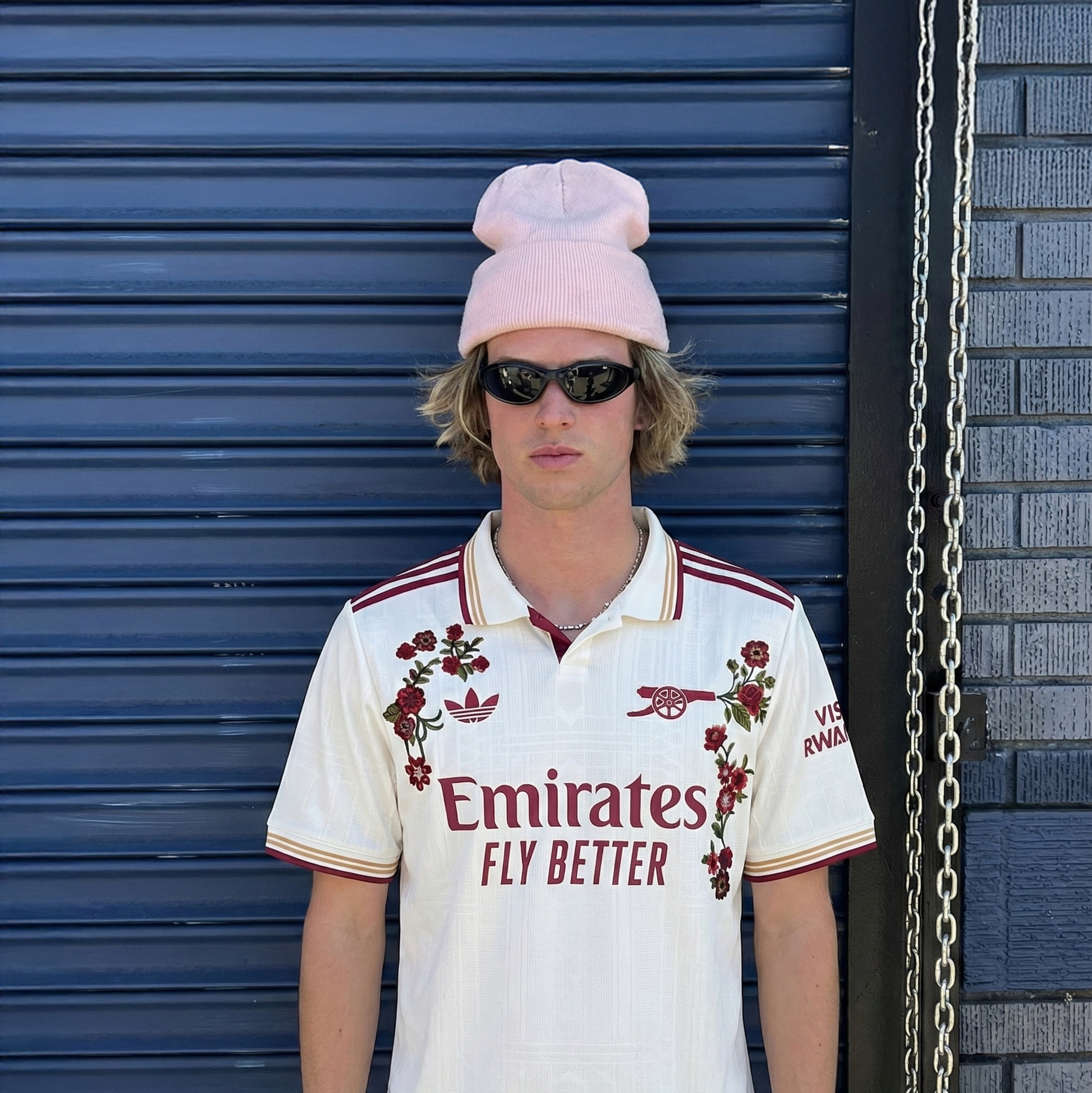 Man wearing Arsenal home football jersey with red floral embroidery, standing against a blue metal shutter in a streetwear style outfit