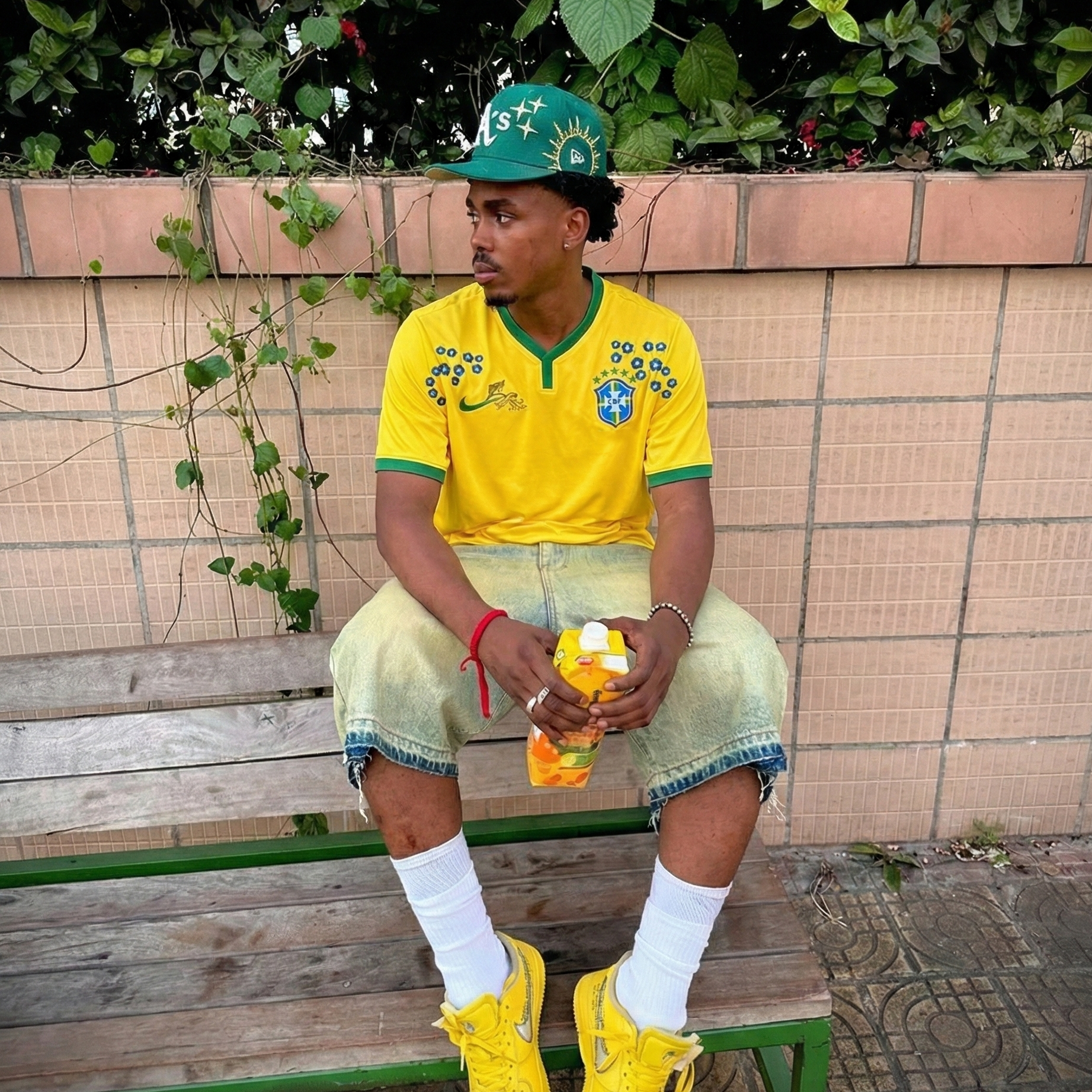 Man wearing a Brazil 2014 home football jersey with floral details, sitting on a bench outdoors, styled with yellow sneakers and casual denim