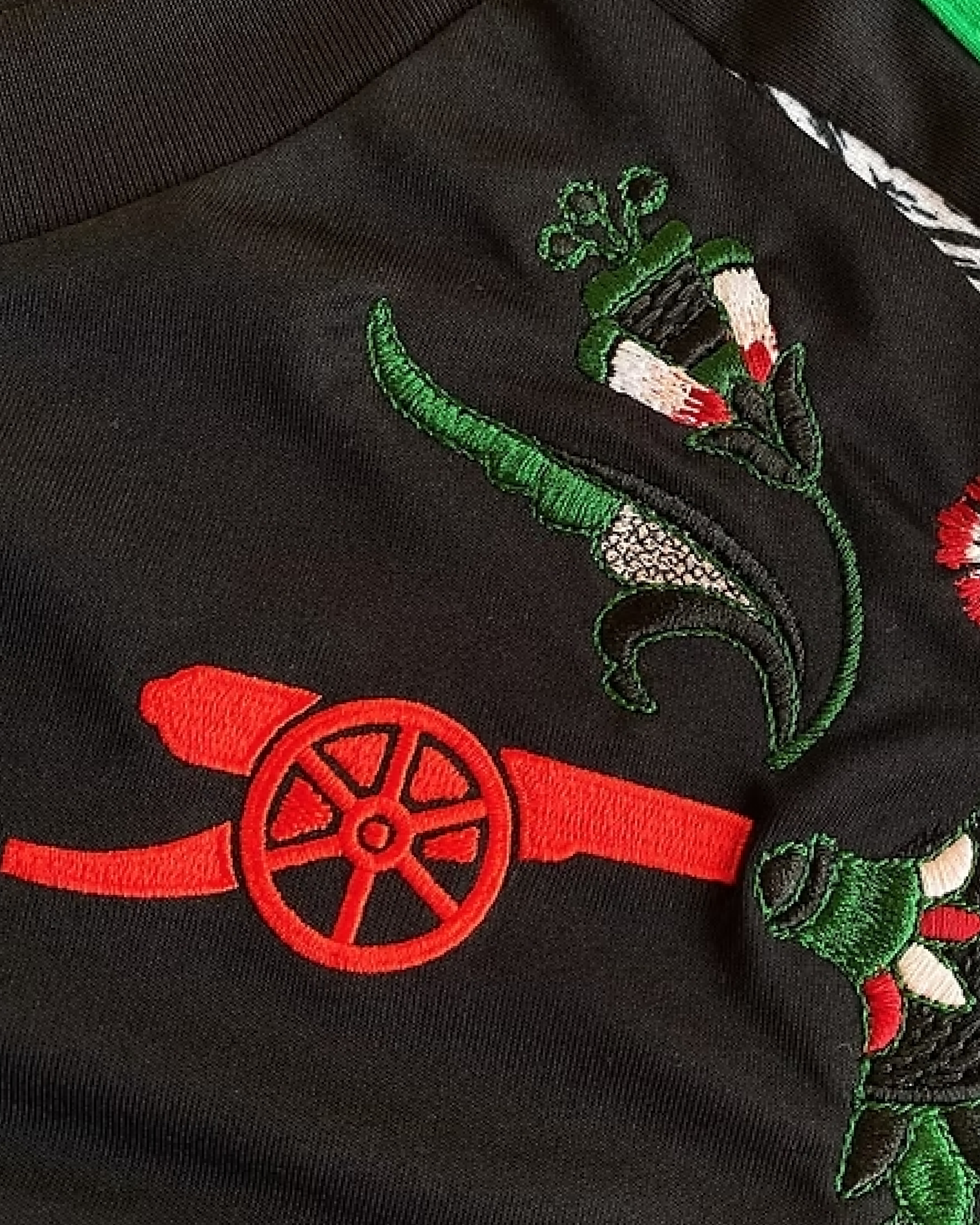 Close-up of the iconic red cannon crest on the Embroidered Arsenal Away Jersey 2024-25, featuring intricate floral stitching and premium black fabric.