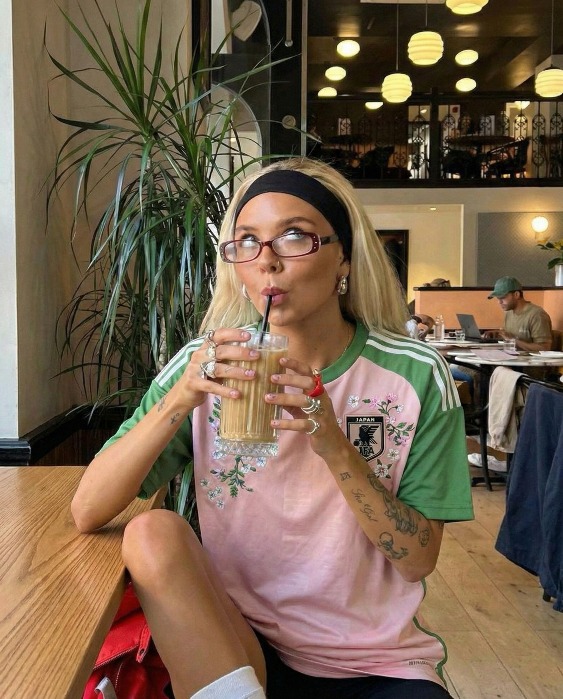 Woman wearing pink and green Japan football jersey with floral embroidery, sitting in a café and drinking an iced coffee, styled in a modern streetwear look