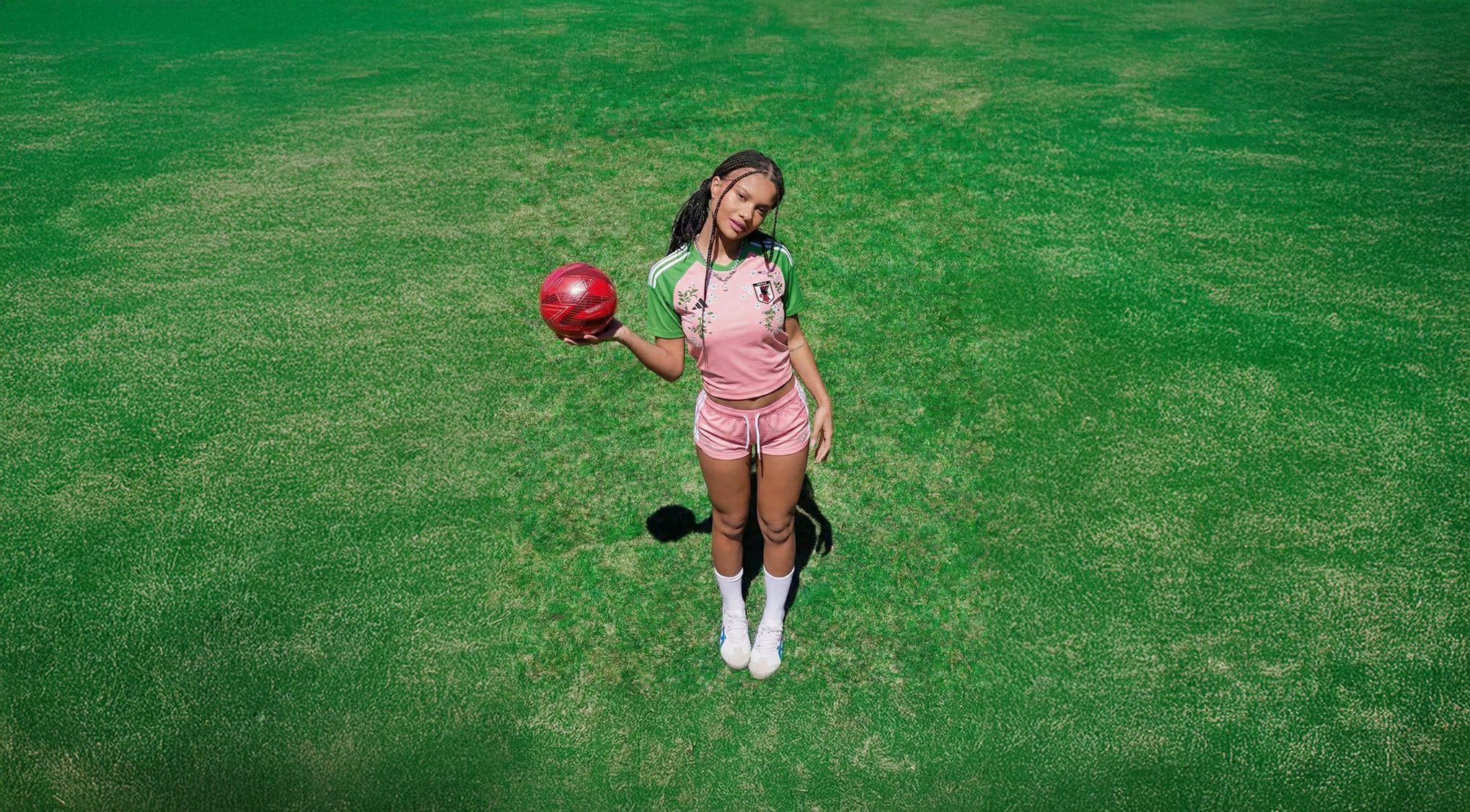 Woman wearing pink and green Japan football jersey with floral embroidery, holding a red football on a green field in a sporty streetwear style