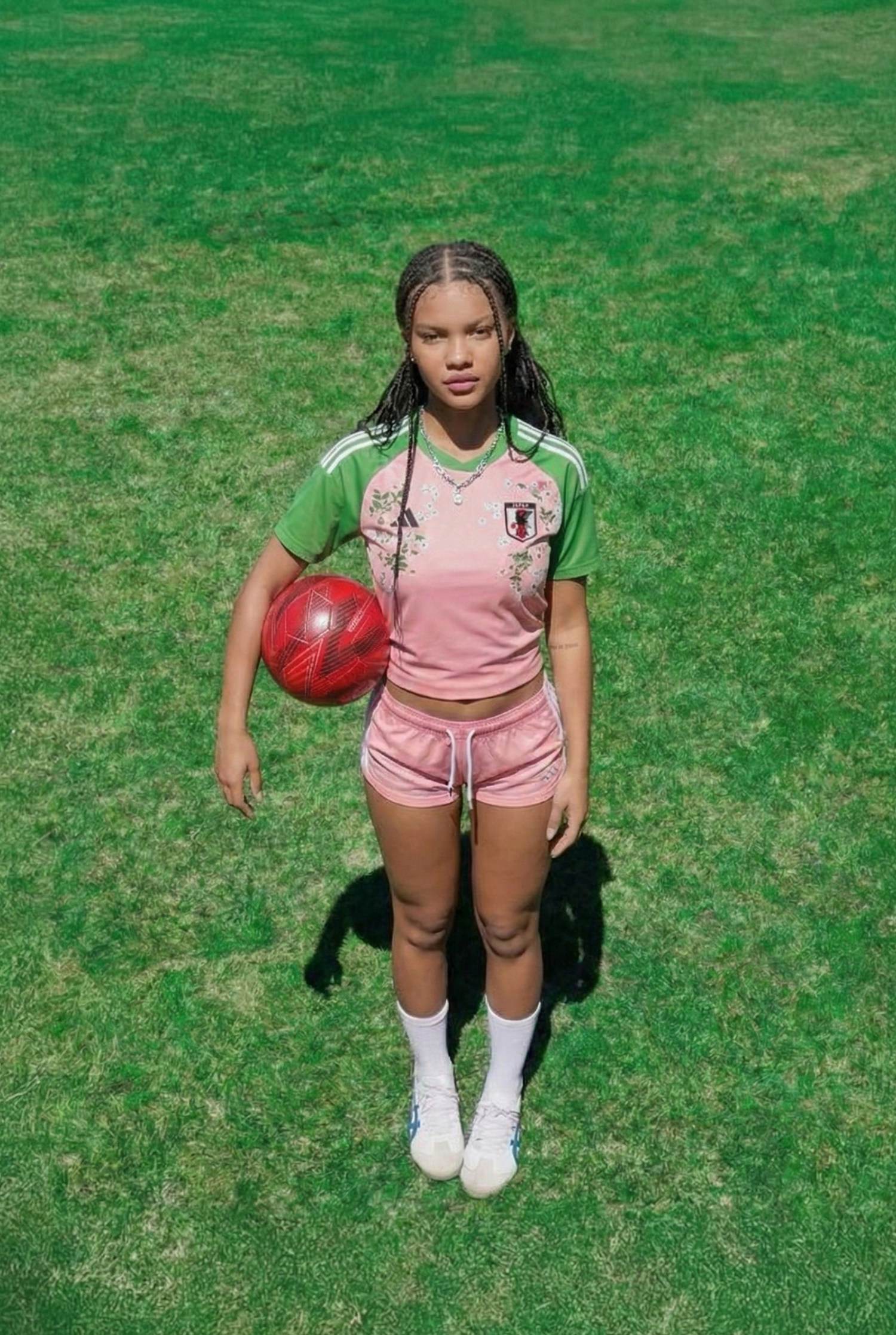 Woman standing on green grass holding a red football while wearing a pink and green Japan football jersey with floral embroidery