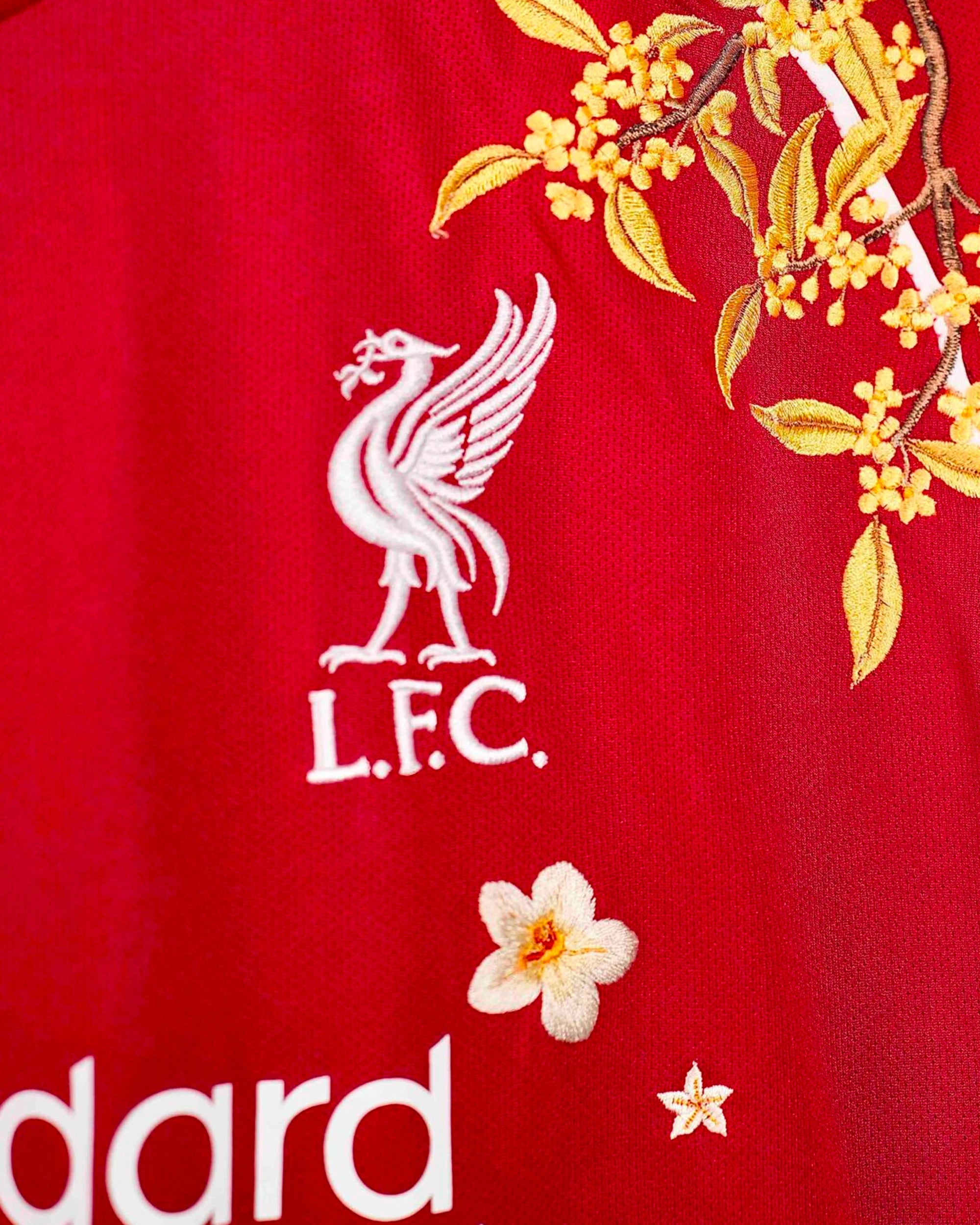 Detailed macro close-up of the Liverpool 'Red Birds' Edition 2025-26 Jersey club crest overlaid with a heavy golden-yellow floral branch.
