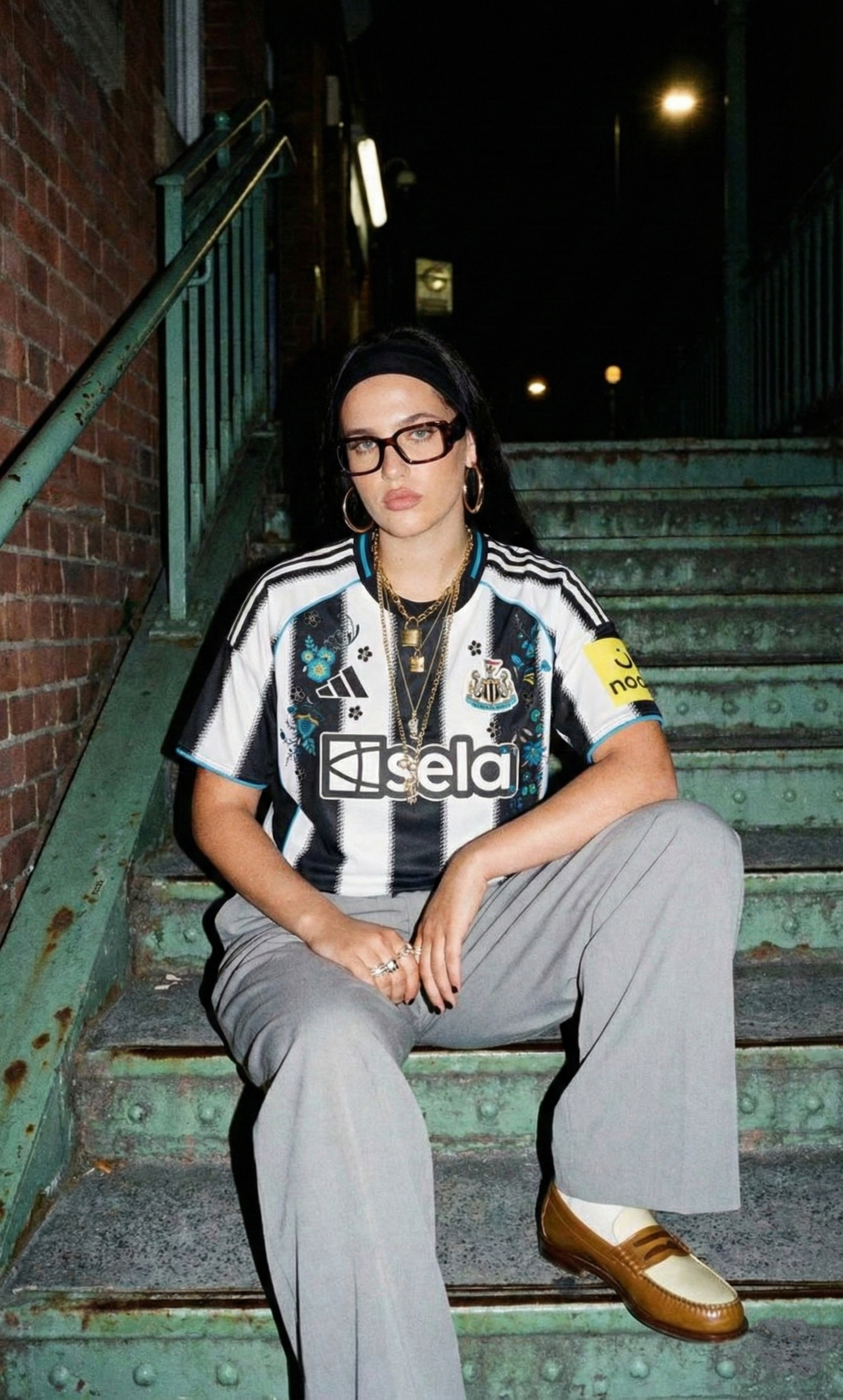Woman wearing a Newcastle United Embroidered football jersey with black and white stripes, styled in a night streetwear look while sitting on outdoor stairs