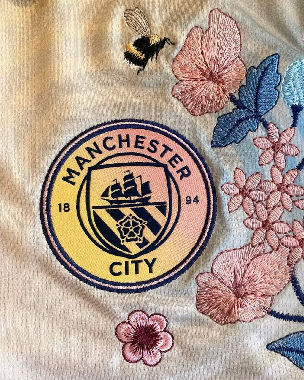 Close-up of the Manchester City crest on the Embroidered Manchester City Fourth Jersey 2024-25, accented by detailed pink flower stitching and a bumblebee motif.